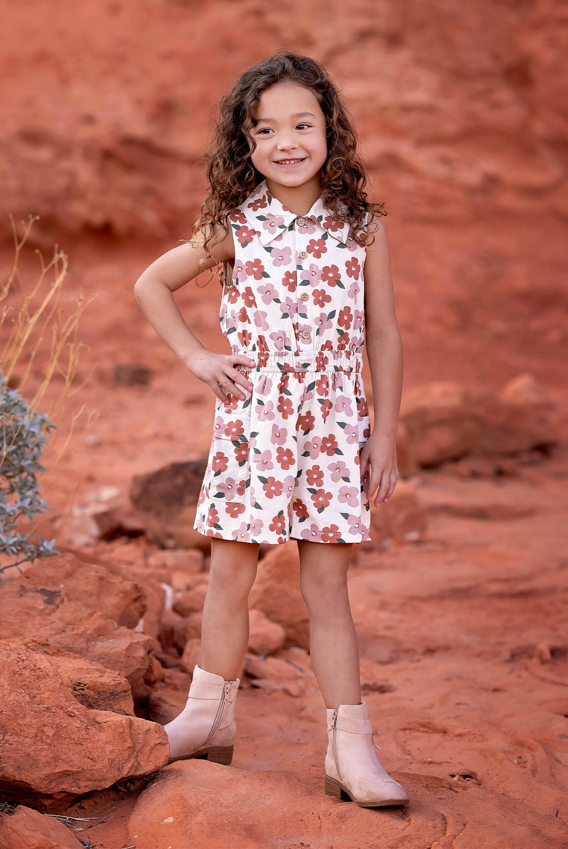 A young girl with curly hair smiles on red rocks, wearing the Mavery Floral Romper by Mabel and Honey with light tan ankle boots, set against a desert landscape of reddish soil and sparse vegetation.