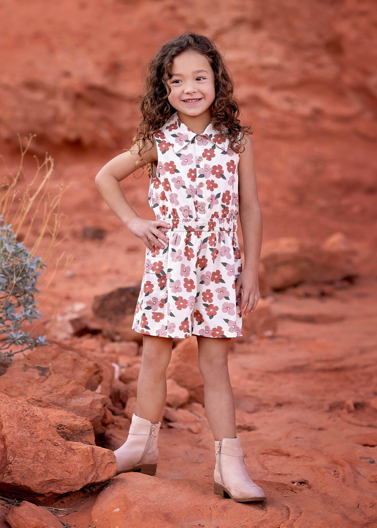 A young girl with curly brown hair smiles while standing on red rocks in a desert landscape, wearing the Mavery Floral Romper by Mabel + Honey and light-colored ankle boots.