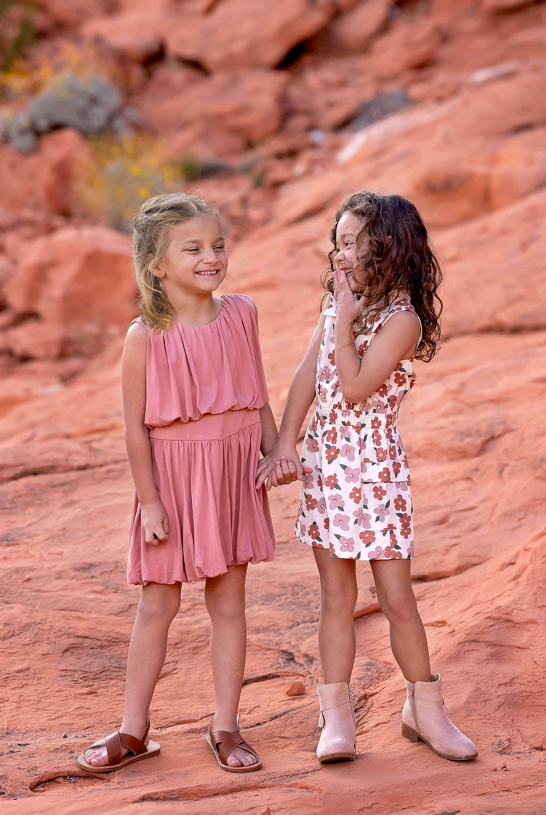 Two young girls stand on reddish rocks, smiling and holding hands. One wears a pink dress and brown sandals; the other is in a Mavery Floral Romper by Mabel and Honey with beige boots. They look happy and playful outdoors.