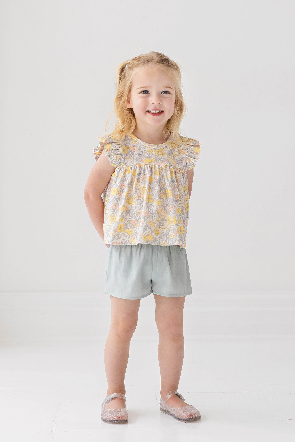 A young blonde girl smiles in the Mila Two Piece Set by Mabel and Honey, featuring a pastel-print top and sage green shorts, paired with sparkly shoes against a plain white background.