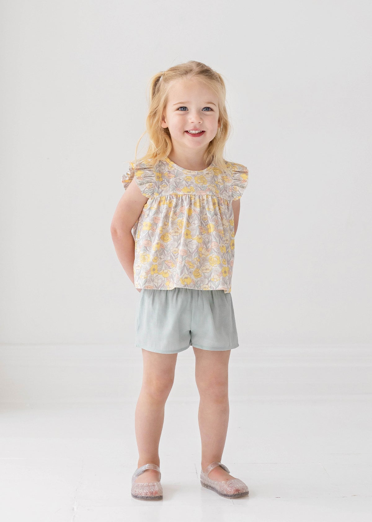A young blonde girl smiles in the Mila Two Piece Set by Mabel and Honey, featuring a pastel-print top and sage green shorts, paired with sparkly shoes against a plain white background.