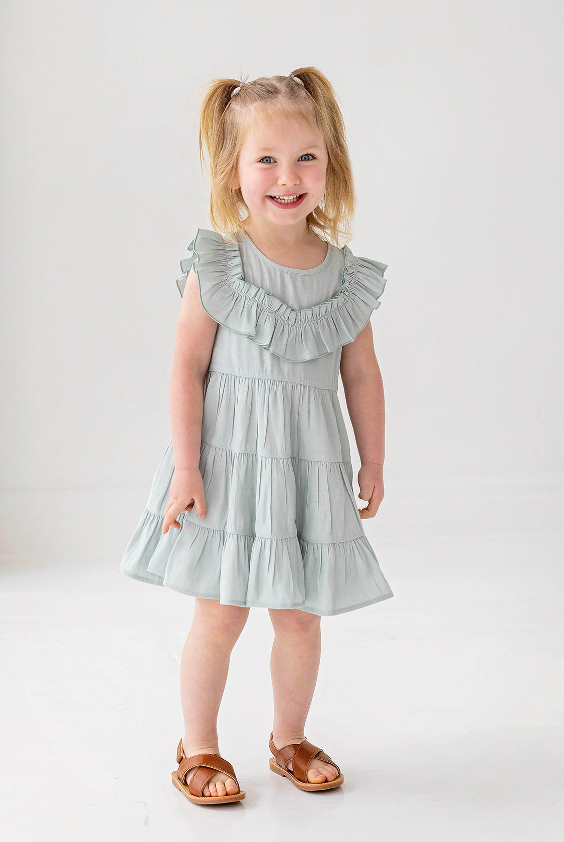 A young child with blonde pigtails smiles while standing against a white background, wearing the Mabel and Honey Mila Green Emerald Dress.