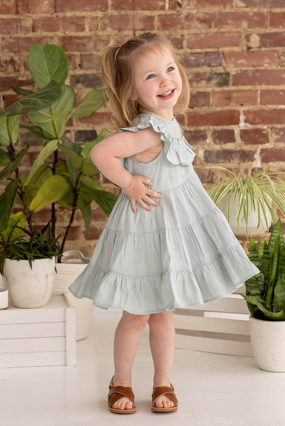 A young girl with blonde pigtails smiles while posing indoors in the Mila Green Emerald Dress by Mabel and Honey, paired with brown sandals near green potted plants and a brick wall.