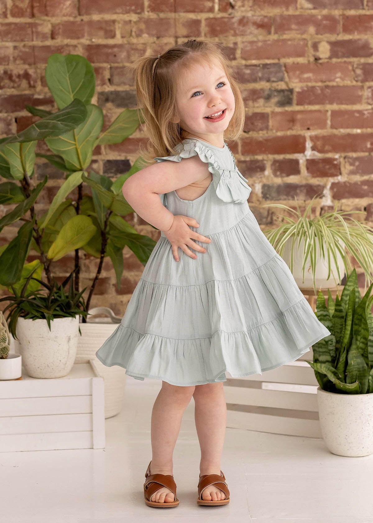 A young girl with blonde pigtails smiles while posing indoors in the Mila Green Emerald Dress by Mabel and Honey, paired with brown sandals near green potted plants and a brick wall.