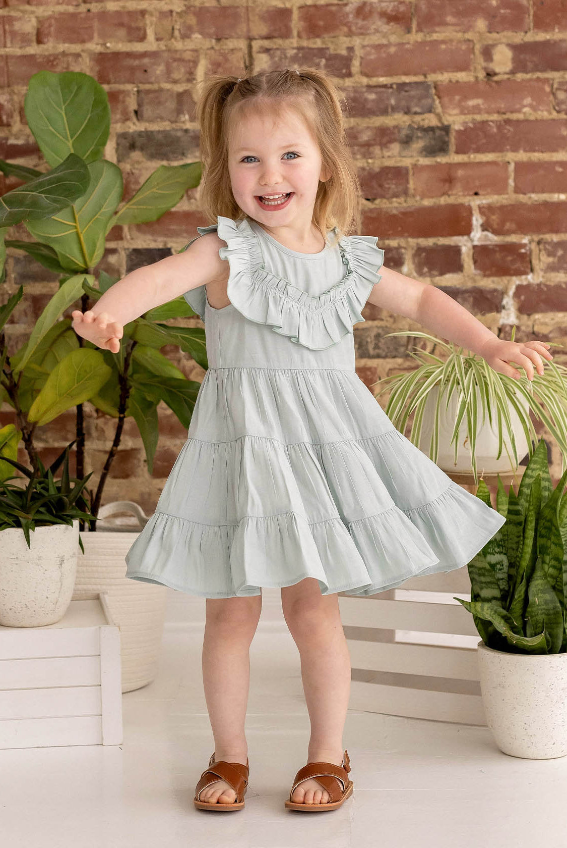 A smiling young girl with light brown pigtails wears the Mila Green Emerald Dress by Mabel and Honey, standing with arms outstretched among potted green plants against a rustic brick wall background, paired with brown sandals.
