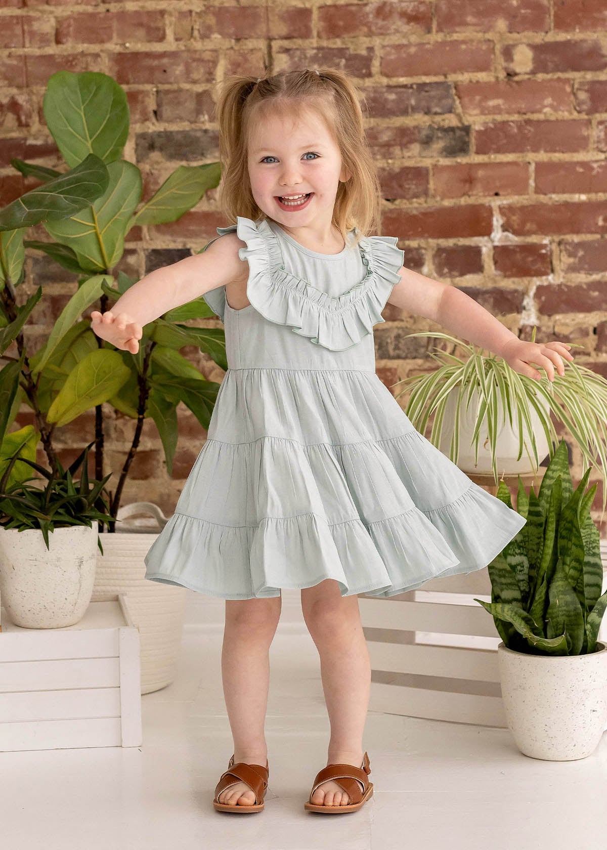 A smiling young girl with light brown pigtails wears the Mila Green Emerald Dress by Mabel and Honey, standing with arms outstretched among potted green plants against a rustic brick wall background, paired with brown sandals.
