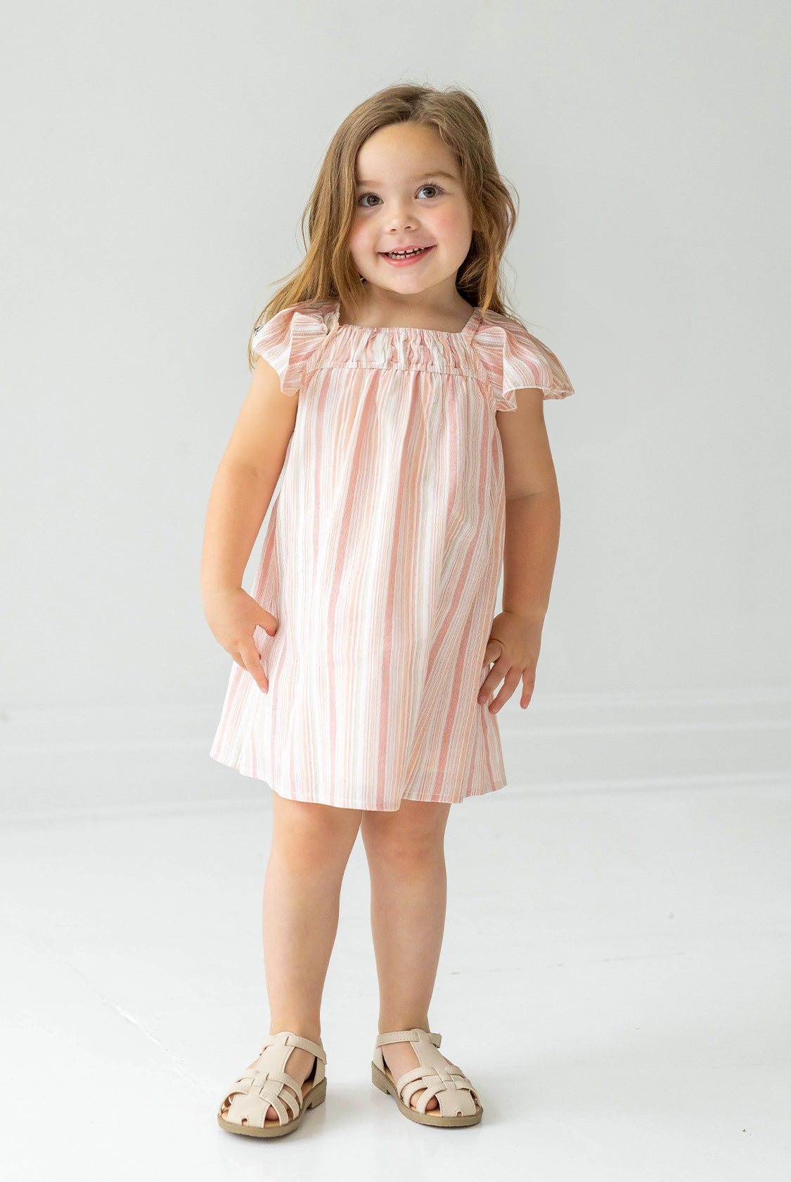 A young girl with long brown hair wears the Sandy Dress by Mabel and Honey, a light pink and white striped dress with flutter sleeves, paired with beige sandals, standing and smiling against a plain white background.