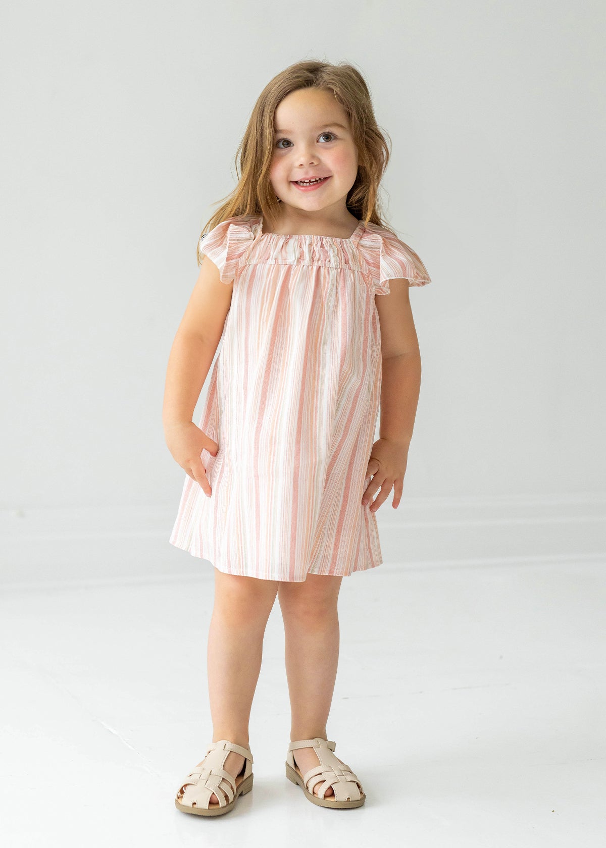 A young girl with long brown hair wears the Sandy Dress by Mabel and Honey, a light pink and white striped dress with flutter sleeves, paired with beige sandals, standing and smiling against a plain white background.