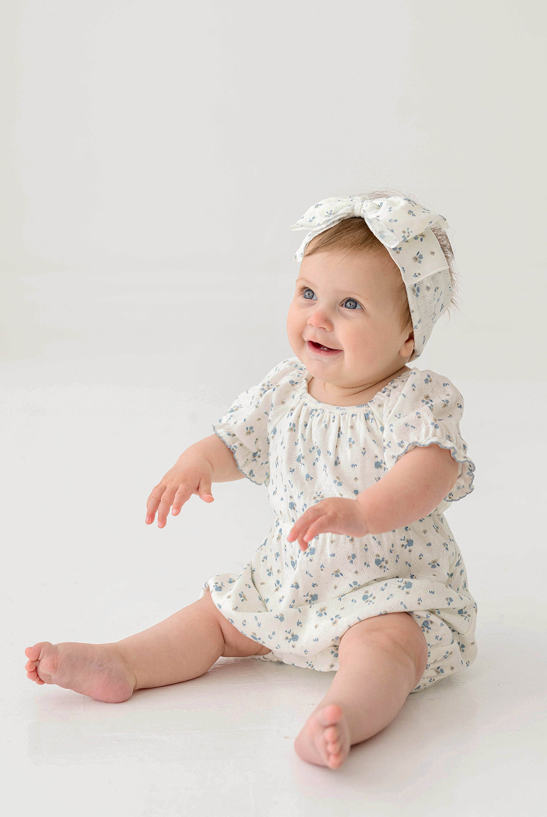 A smiling baby wears the Bluebell Baby Romper from Mabel and Honey with puff sleeves and a matching headband, sitting on the floor against a plain white background.