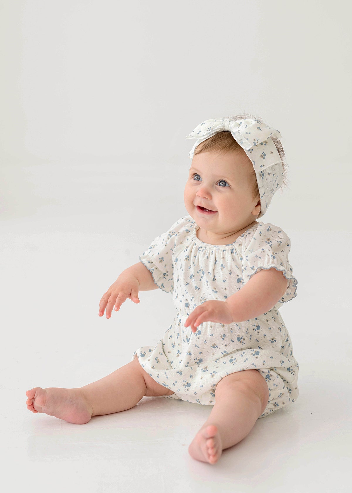 A smiling baby wears the Bluebell Baby Romper from Mabel and Honey with puff sleeves and a matching headband, sitting on the floor against a plain white background.