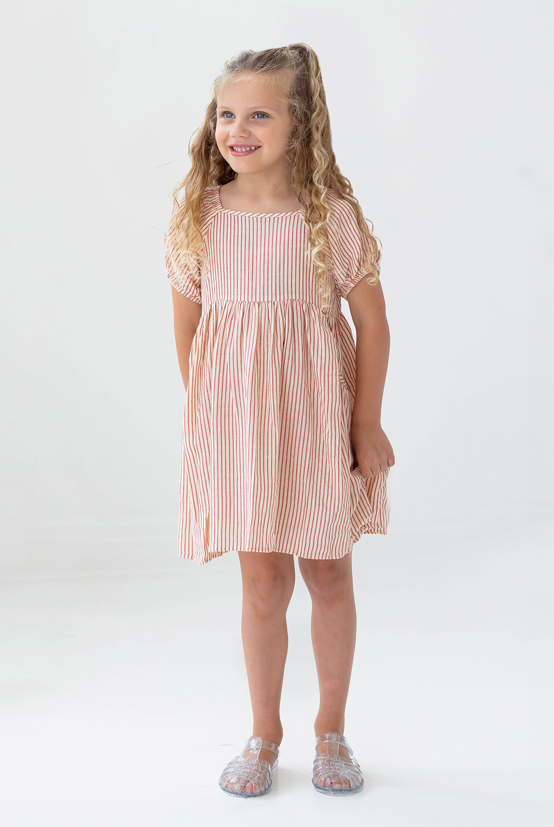 A young girl with long curly blonde hair smiles in the Mabel and Honey Cranberry Crush Dress—a peach and white striped, short-sleeve girls dress—paired with transparent sandals against a plain white background.