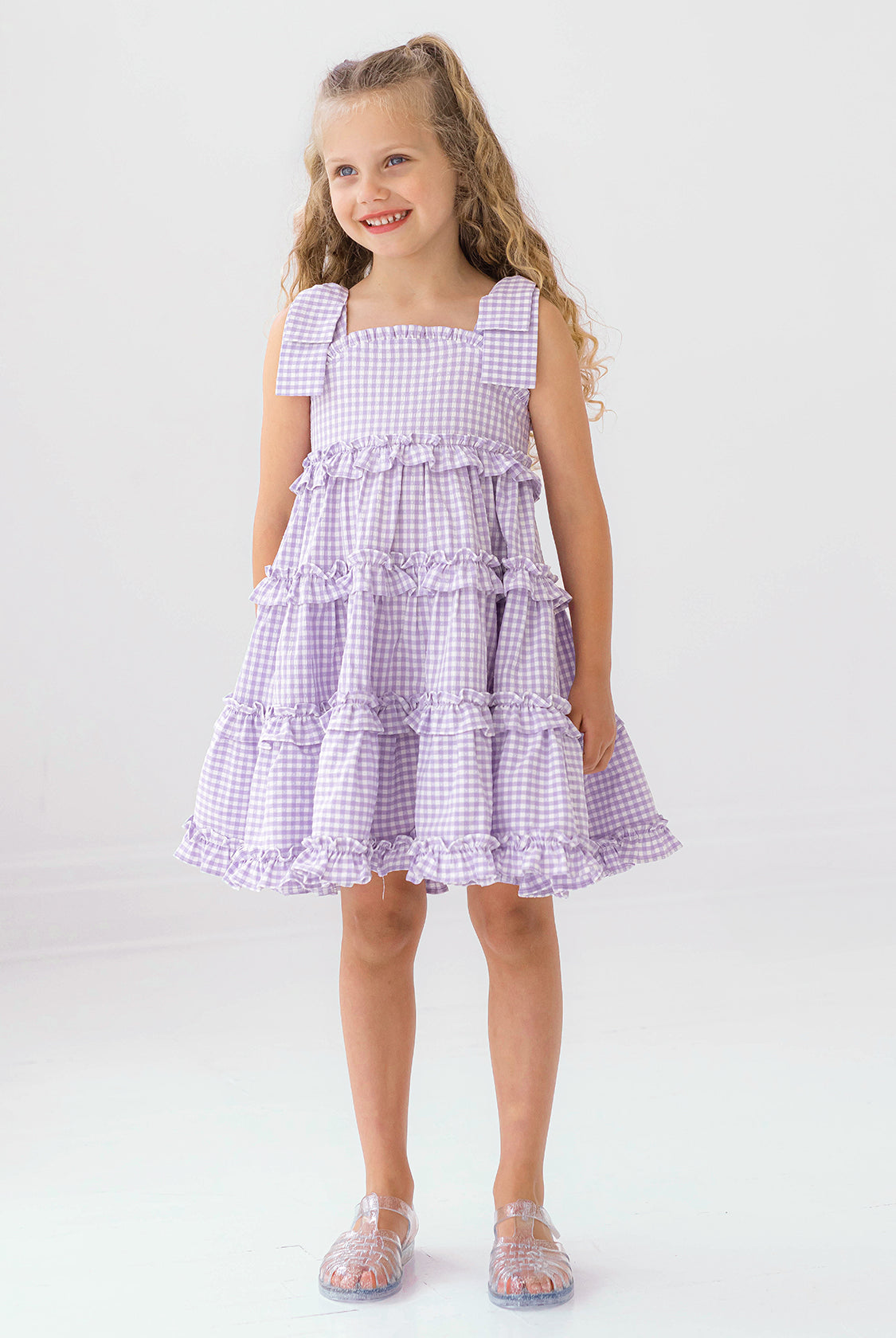 A young girl with long curly hair smiles in the Mabel and Honey Super Bloom Tiered Dress, featuring lavender gingham, ruffles, and bows. She pairs it with clear sandals while standing against a plain white background.