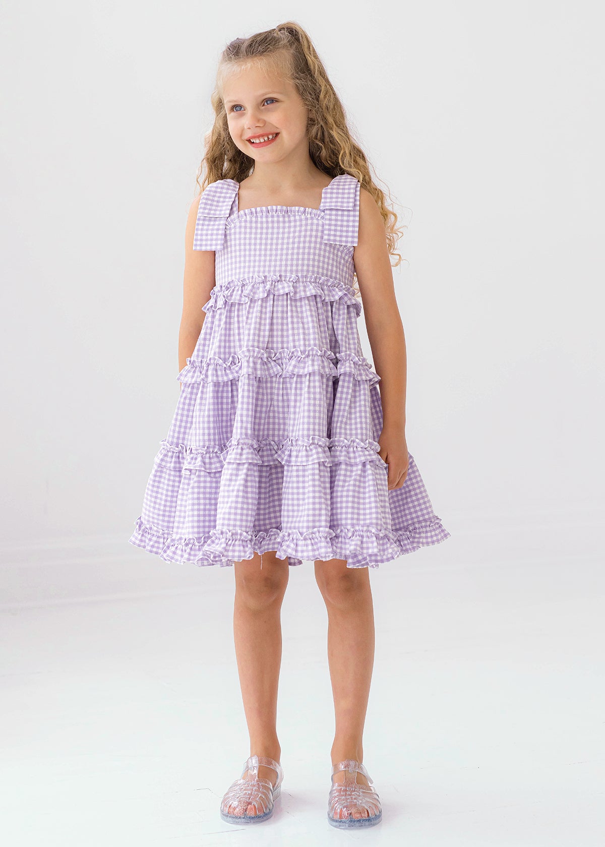 A young girl with long curly hair smiles in the Mabel and Honey Super Bloom Tiered Dress, featuring lavender gingham, ruffles, and bows. She pairs it with clear sandals while standing against a plain white background.