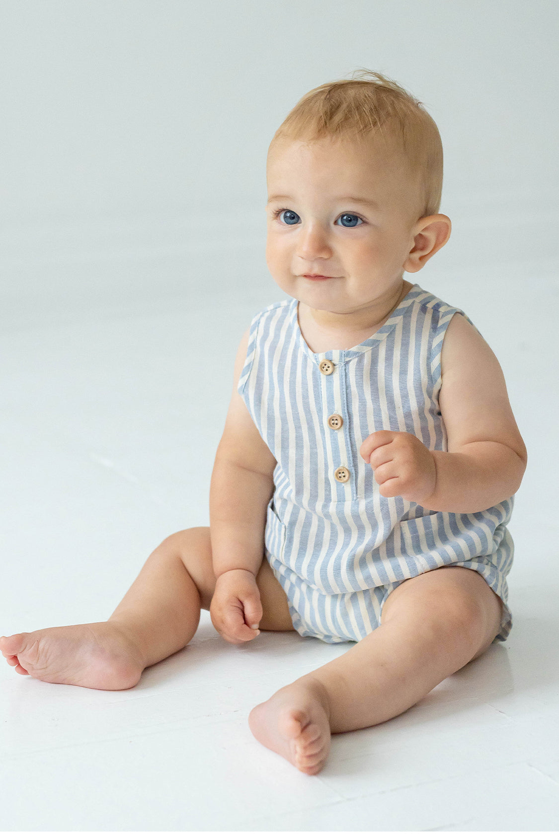 A baby with light hair and blue eyes sits on a white floor, wearing the Beckett and Bear Blake Baby Romper in light blue stripes with wooden buttons. The baby looks slightly upward and smiles softly.