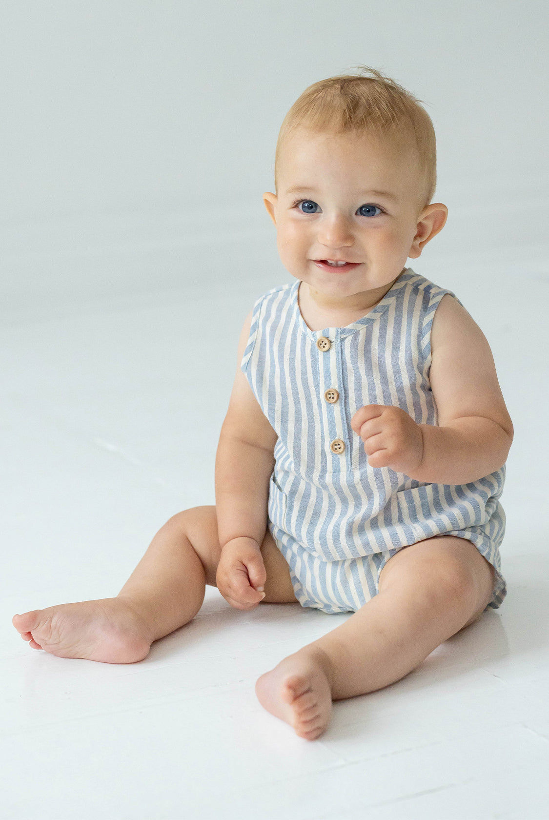 A smiling baby with short light hair sits on the floor in the Beckett and Bear Blake Baby Romper, featuring light blue stripes and wooden buttons, looking slightly to the side.