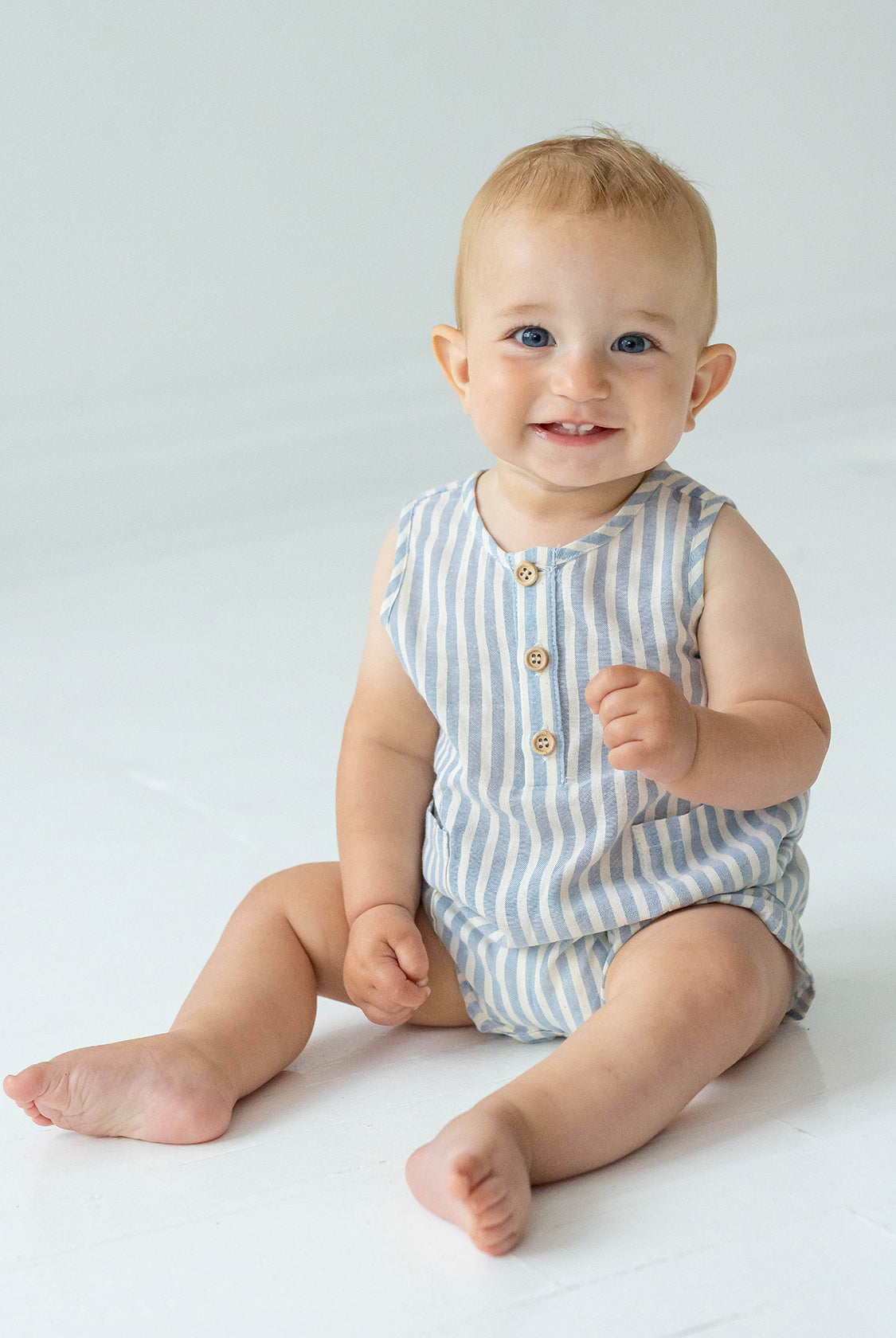 A smiling baby with blue eyes and light hair sits barefoot on the floor in a Beckett and Bear Blake Baby Romper, looking at the camera against a plain, light background.