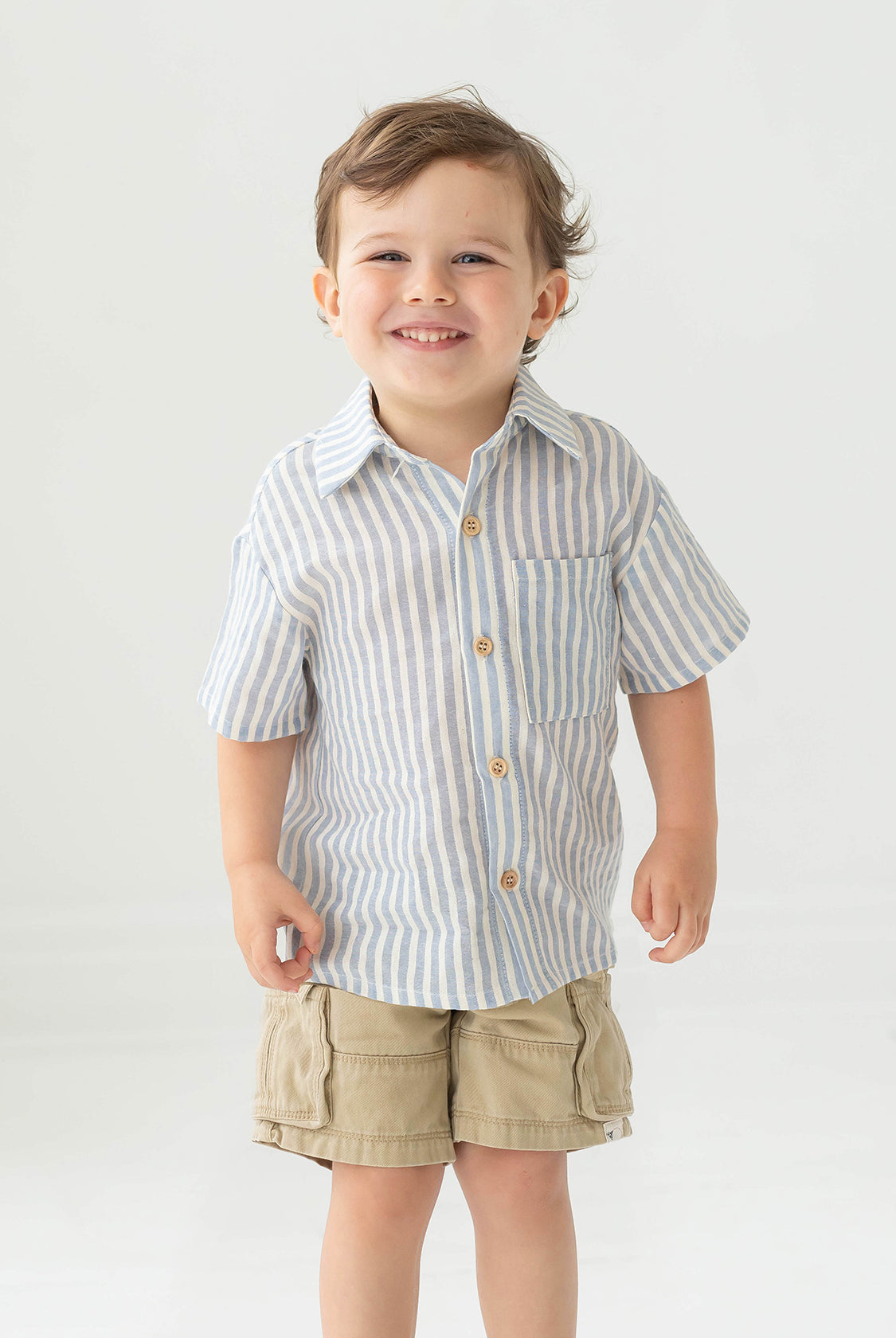 A young boy with short brown hair smiles while wearing the Blake Top from Beckett and Bear, a light blue striped, 100% cotton shirt, paired with beige cargo shorts, standing against a plain light background.