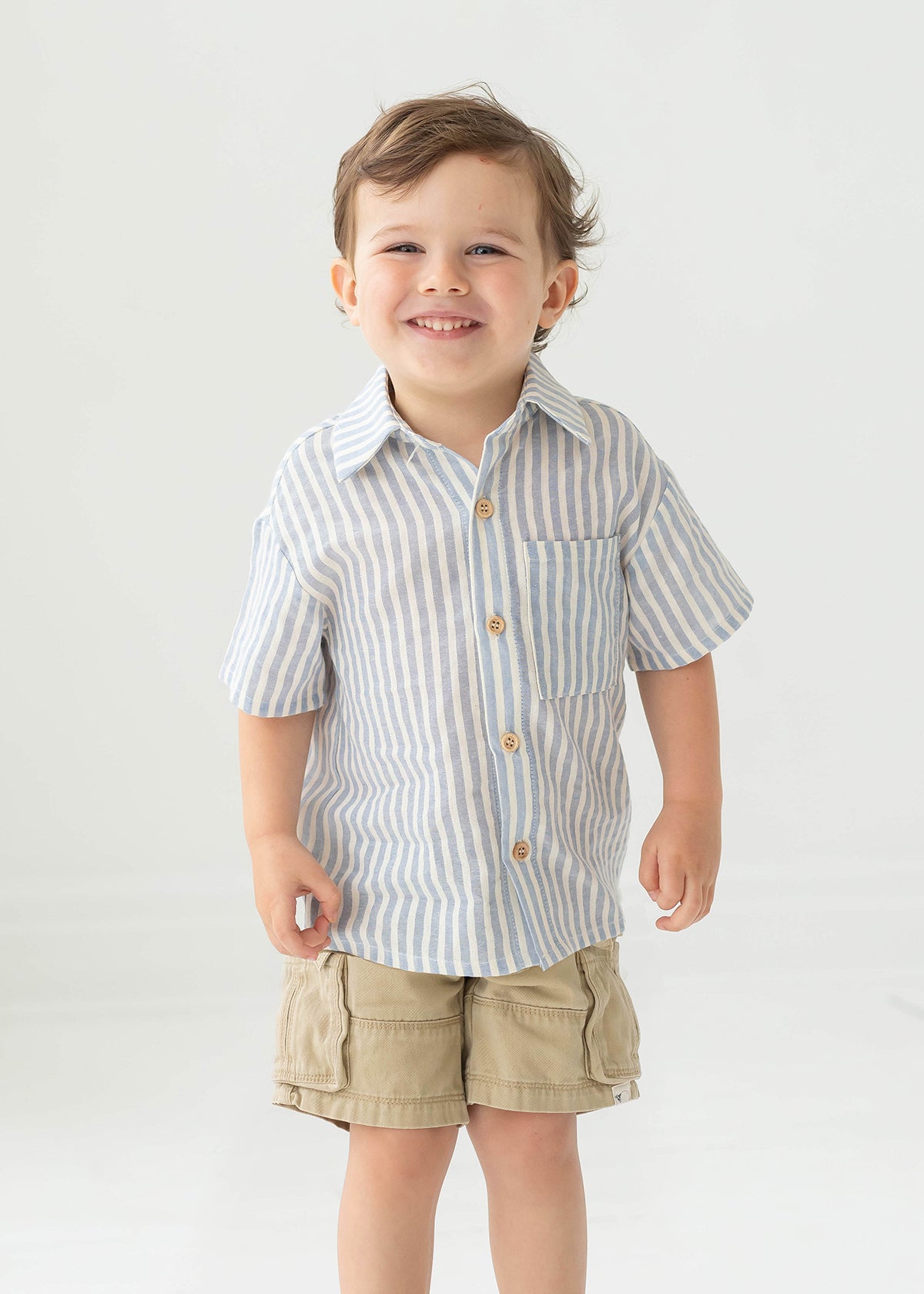 A young boy with short brown hair smiles while wearing the Blake Top from Beckett and Bear, a light blue striped, 100% cotton shirt, paired with beige cargo shorts, standing against a plain light background.