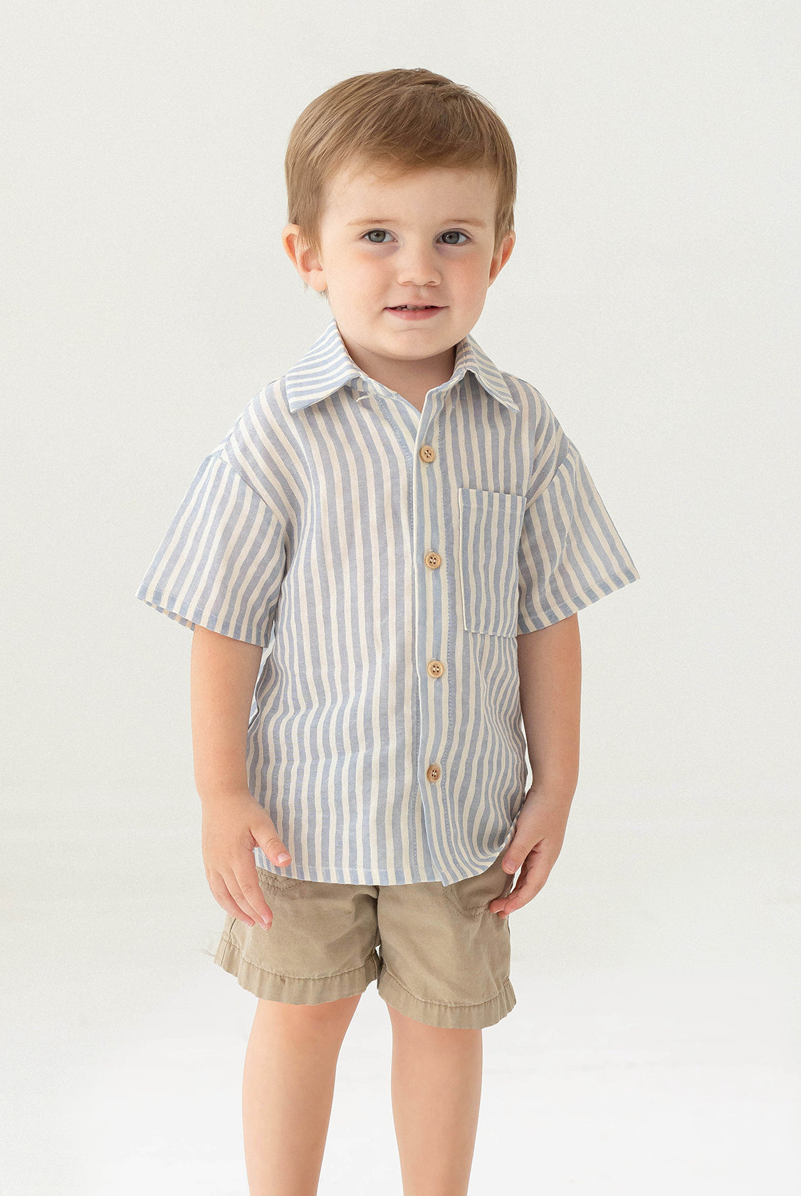 A young boy with short light brown hair stands against a plain background, smiling slightly in the Beckett and Bear Blake Top, a 100% cotton light blue striped short-sleeve button-up shirt, paired with beige shorts.