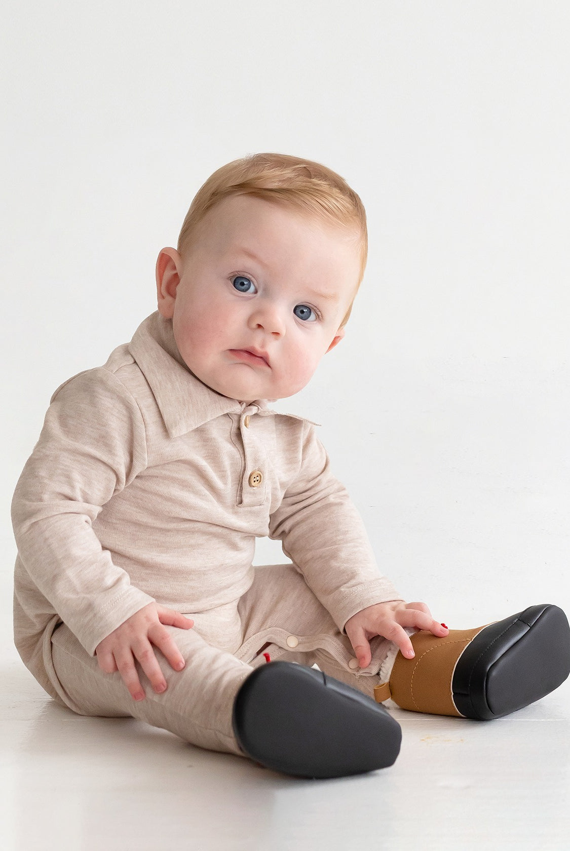 A baby with light hair and blue eyes sits on the floor in a beige Millie Brothers Romper by Beckett and Bear, featuring a collared neckline, long sleeves, and black shoes. The child gazes to the side against a plain white background.