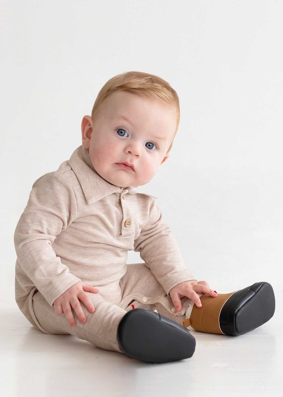 A baby with light hair and blue eyes sits on the floor in a beige Millie Brothers Romper by Beckett and Bear, featuring a collared neckline, long sleeves, and black shoes. The child gazes to the side against a plain white background.