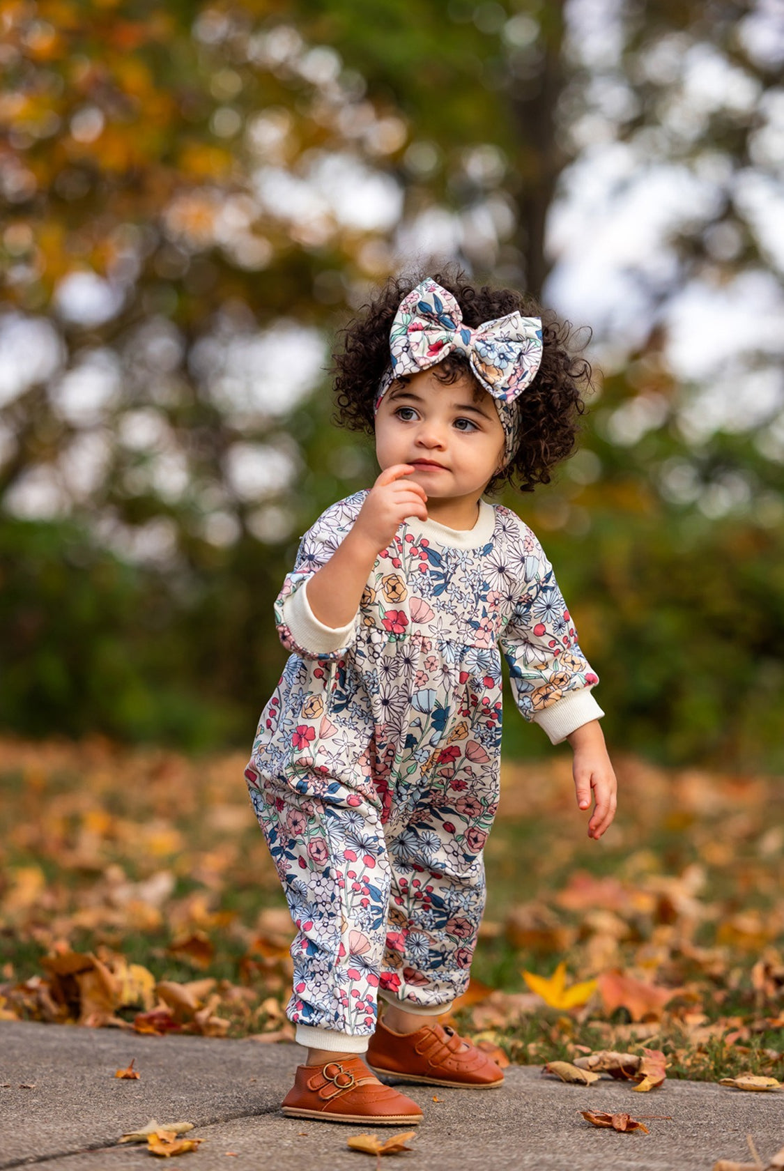 A toddler with curly hair wears the Mabel and Honey Melody Romper and a matching bow headband, standing outdoors on a leaf-strewn path with autumn foliage in the background.
