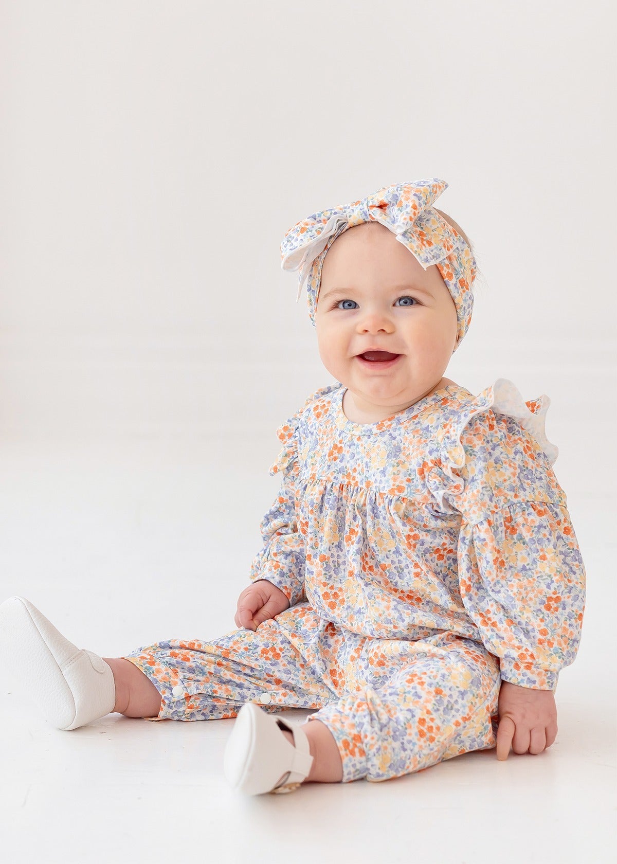 A smiling baby sits on the floor in the Mabel and Honey Darling Orchard Romper with a matching headband and white shoes, against a plain light background.