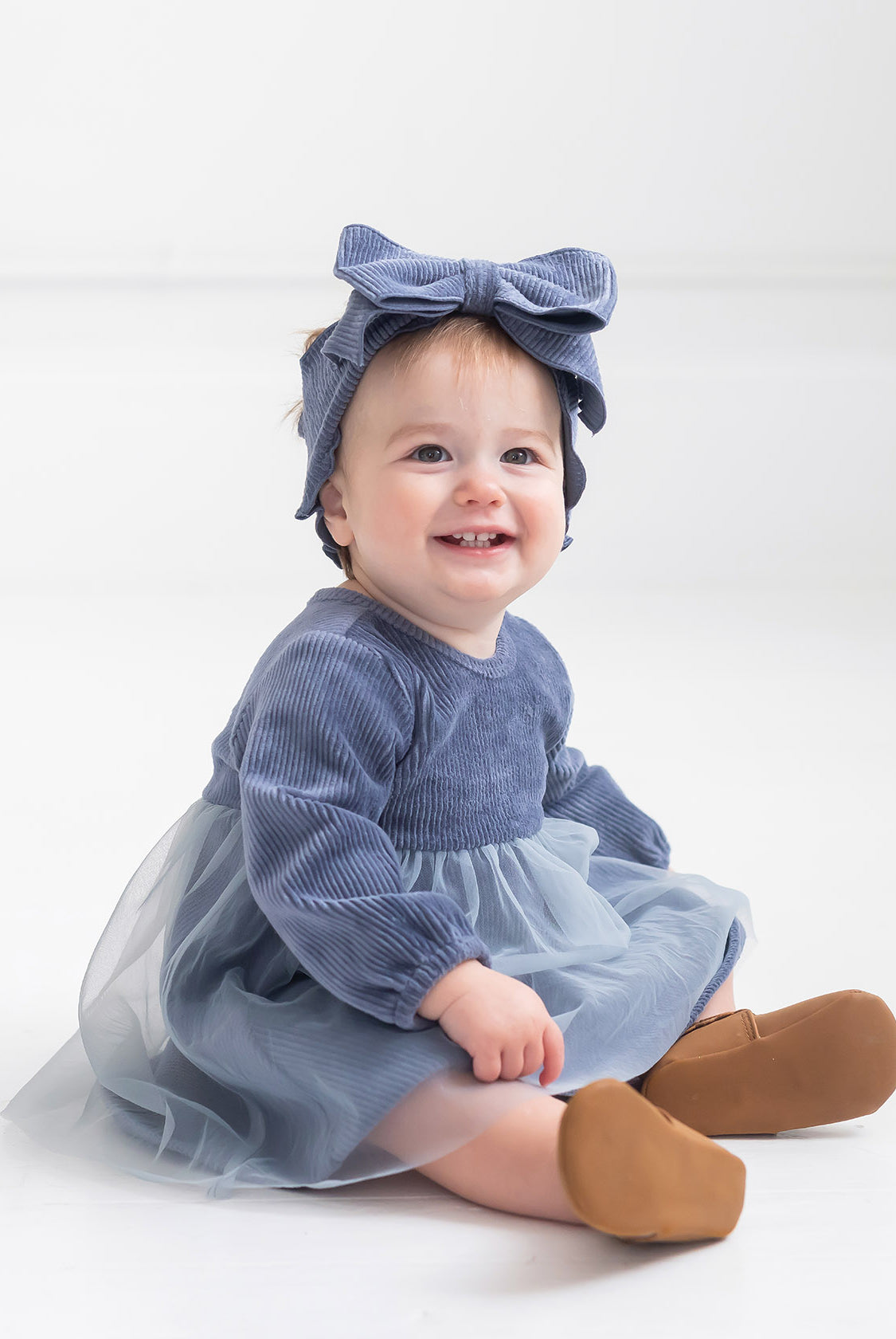 A smiling baby with light hair sits on the floor in a Mabel and Honey Skylar Blue Suede Dress, styled with a tulle skirt, corduroy bodice, a matching blue bow headband, and brown shoes against a plain white background.