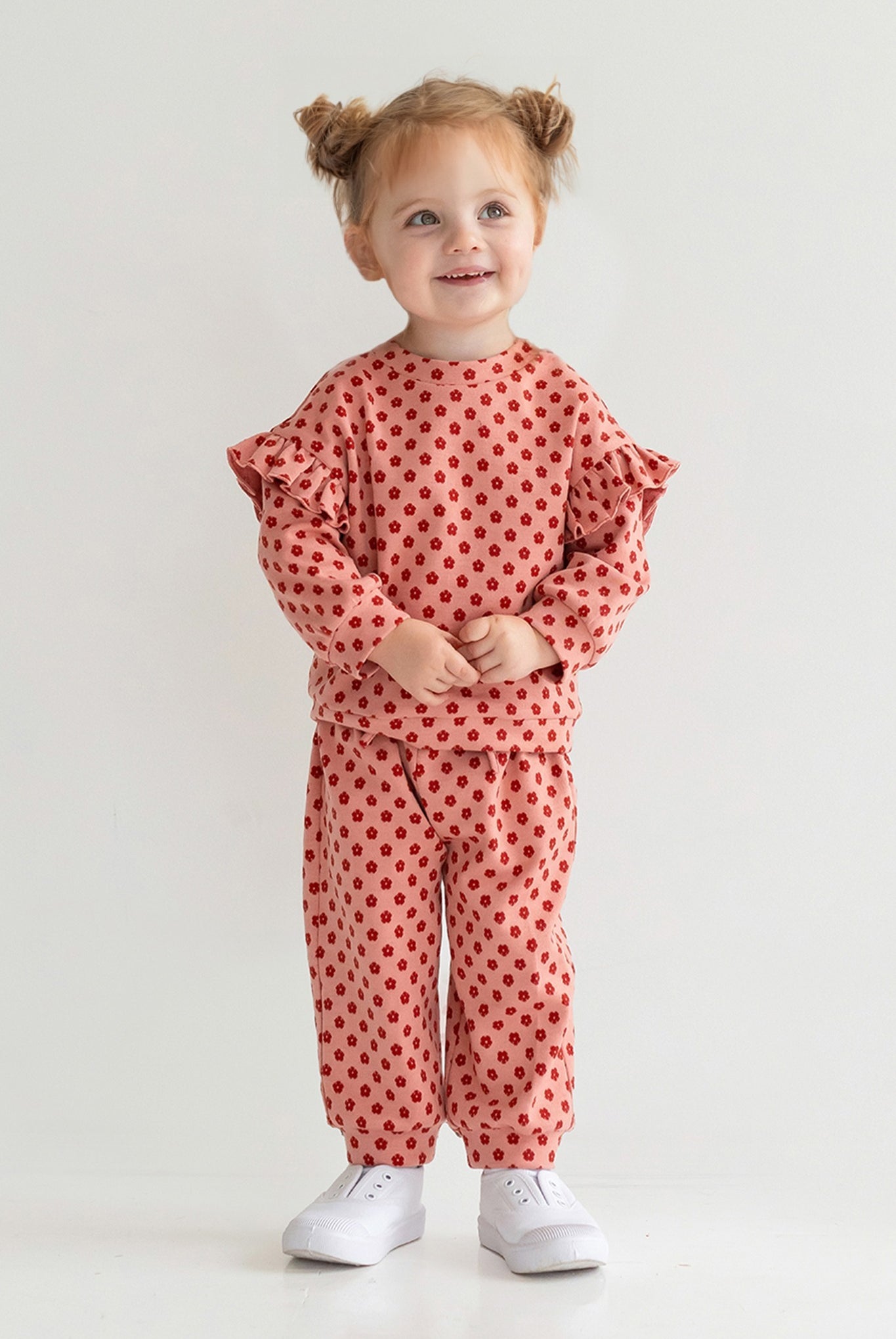 A young child with light brown hair in pigtails smiles while wearing the Mabel and Honey Red Velvet Two Piece Set, featuring a matching knit top and pants, paired with white sneakers against a plain light background.