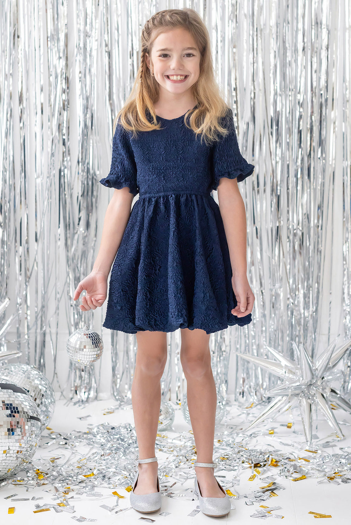 A young girl wears the Isobella and Chloe Nightingale Navy Dress, smiling in front of silver tinsel curtains and holding a small disco ball, surrounded by other disco balls, silver stars, and confetti.