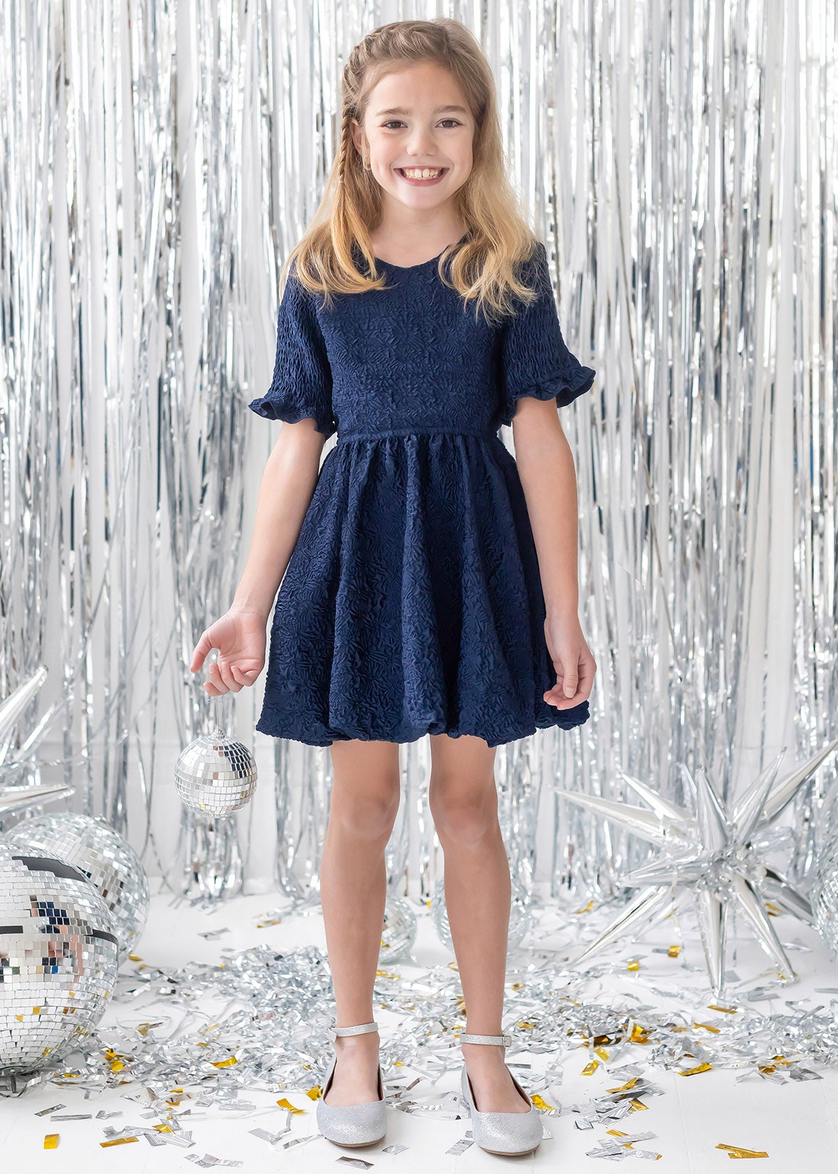 A young girl wears the Isobella and Chloe Nightingale Navy Dress, smiling in front of silver tinsel curtains and holding a small disco ball, surrounded by other disco balls, silver stars, and confetti.