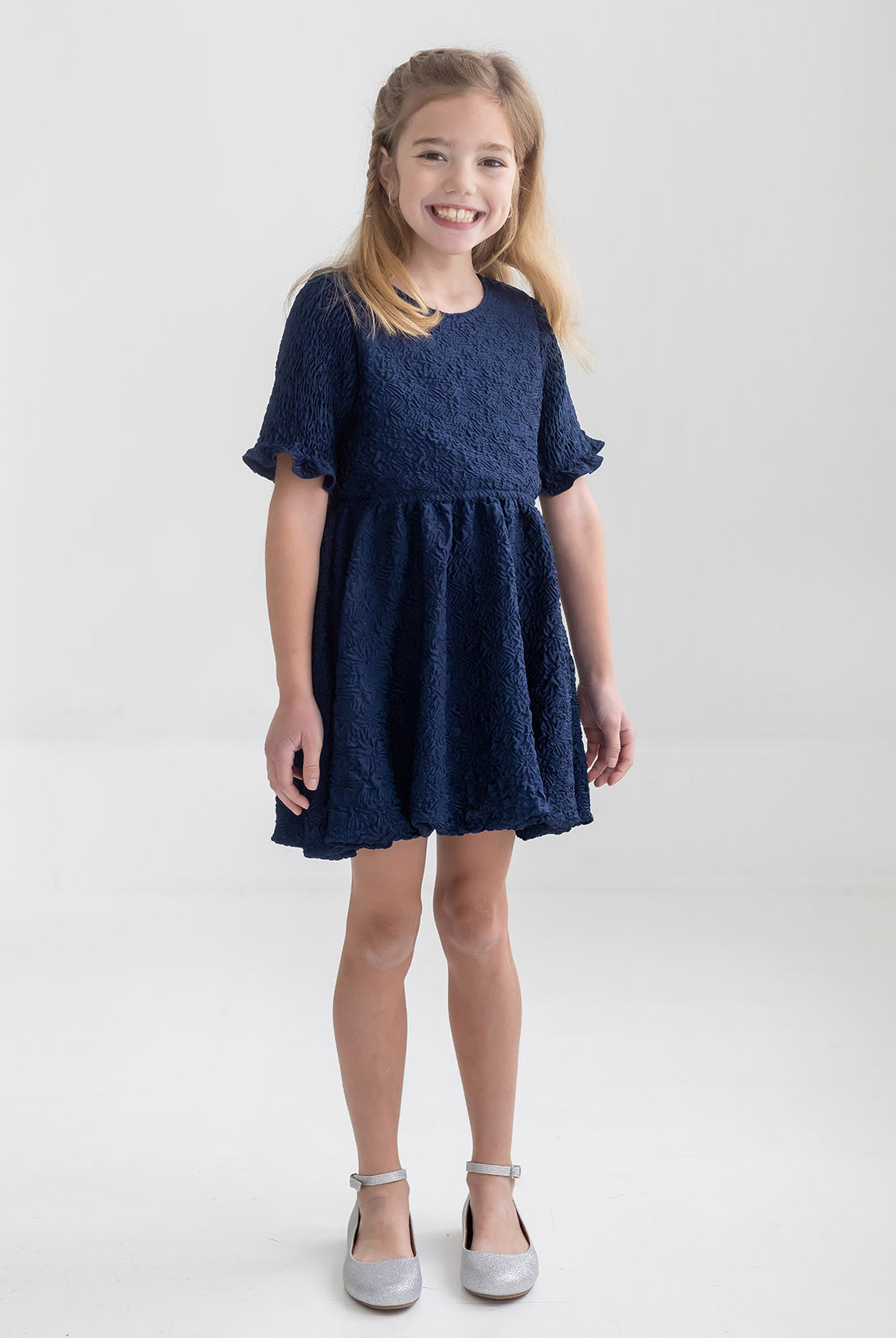 A young girl with light brown hair smiles in a bright, minimal room wearing the Isobella and Chloe Nightingale Navy Dress, made from textured fabric, paired with silver shoes.
