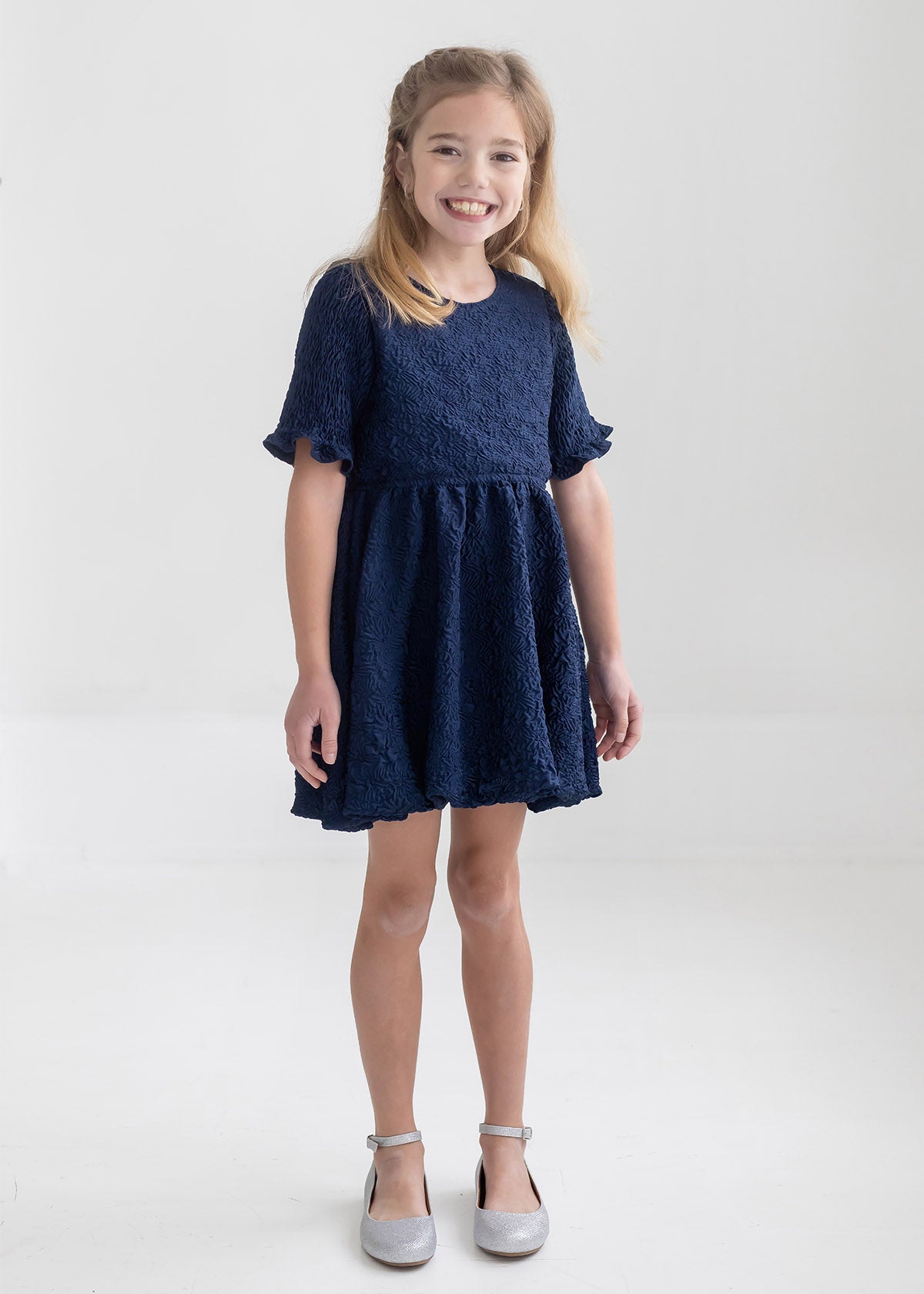 A young girl with light brown hair smiles in a bright, minimal room wearing the Isobella and Chloe Nightingale Navy Dress, made from textured fabric, paired with silver shoes.