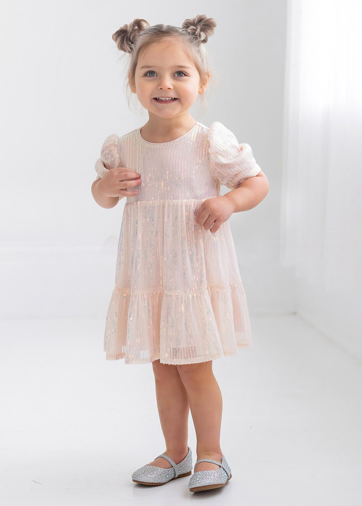 A young girl with light brown hair in two buns smiles while wearing the Isobella and Chloe Taylor Sequined Little Big Girl Dress, featuring puffy sleeves, in a bright white room with silver shoes.