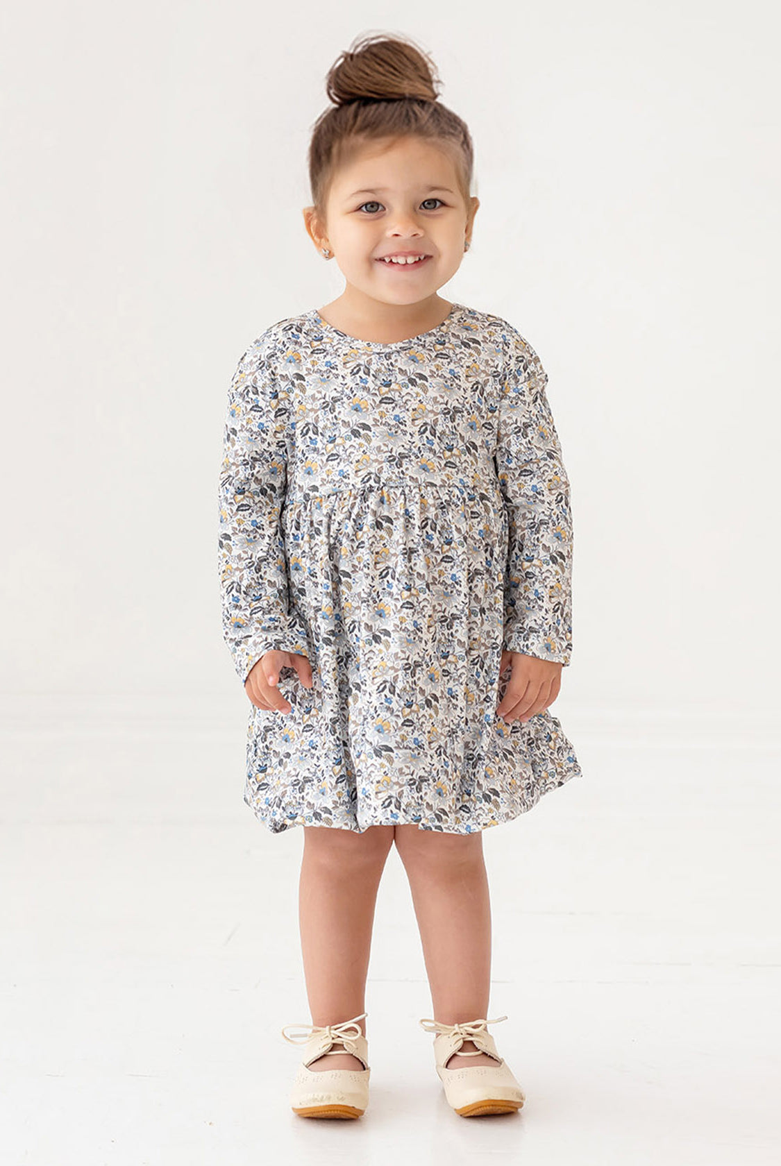 A young girl with brown hair styled in a top bun smiles while standing. She wears the Mabel and Honey Hazelnut Dress, a girls floral long-sleeve dress in light blue, beige, and cream, with beige shoes against a white background.