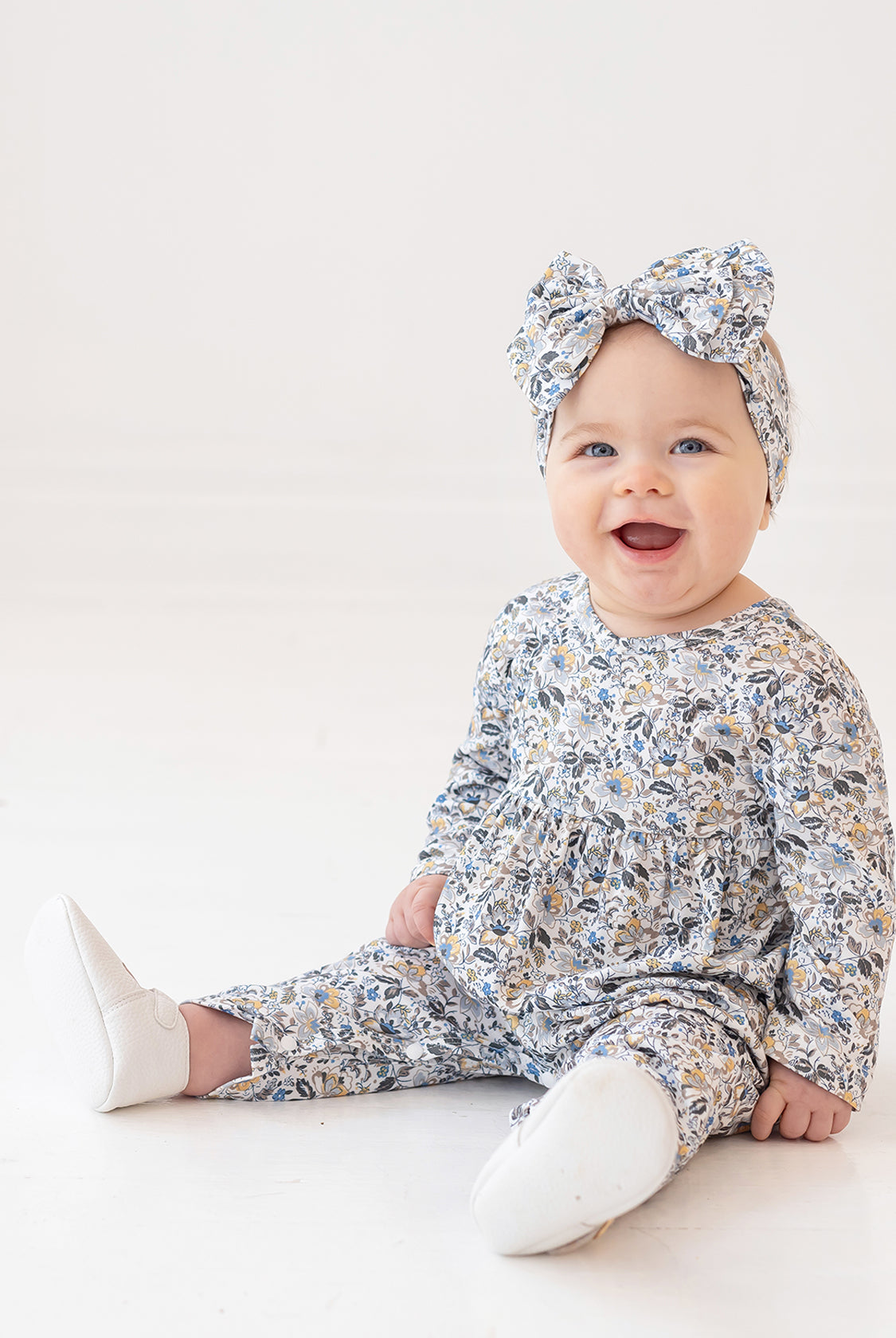 A smiling baby girl wears the Hazelnut Romper by Mabel and Honey with a matching headband, sitting on a white floor against a plain white background.