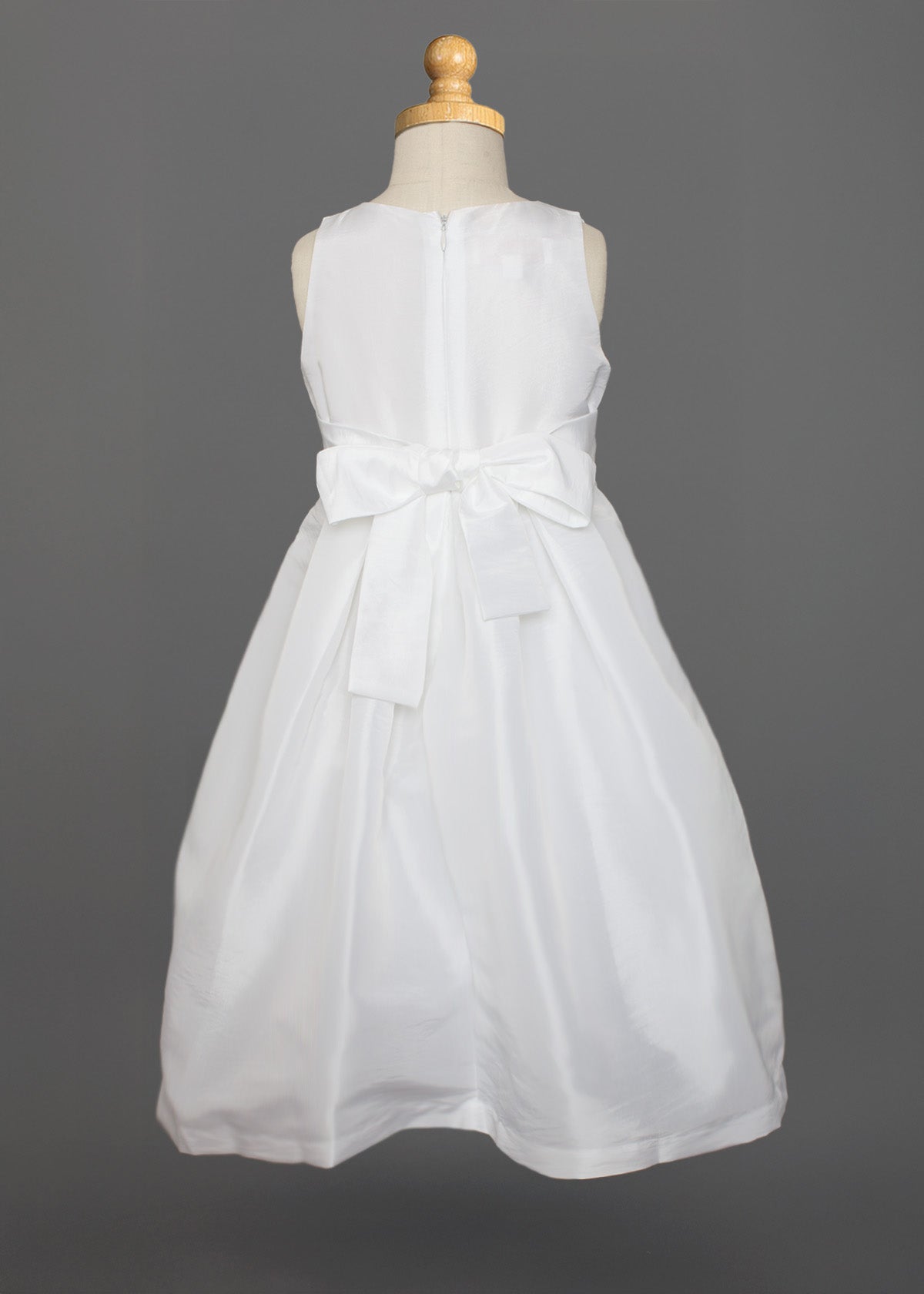The Mabel and Honey Taffeta Bow Communion Dress features a sleeveless white design, full taffeta skirt, large back bow, and a beaded pearl neckline, displayed on a dress form against a gray background.