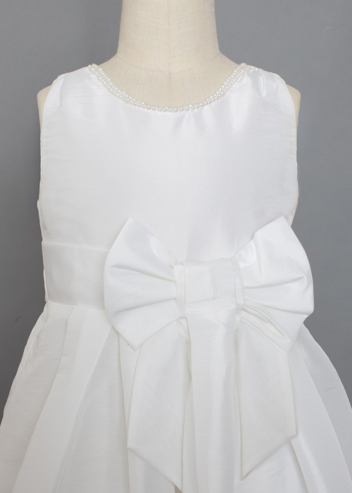 The Mabel and Honey Taffeta Bow Communion Dress features a beaded pearl neckline and a large decorative waist bow, displayed on a mannequin against a plain gray background.
