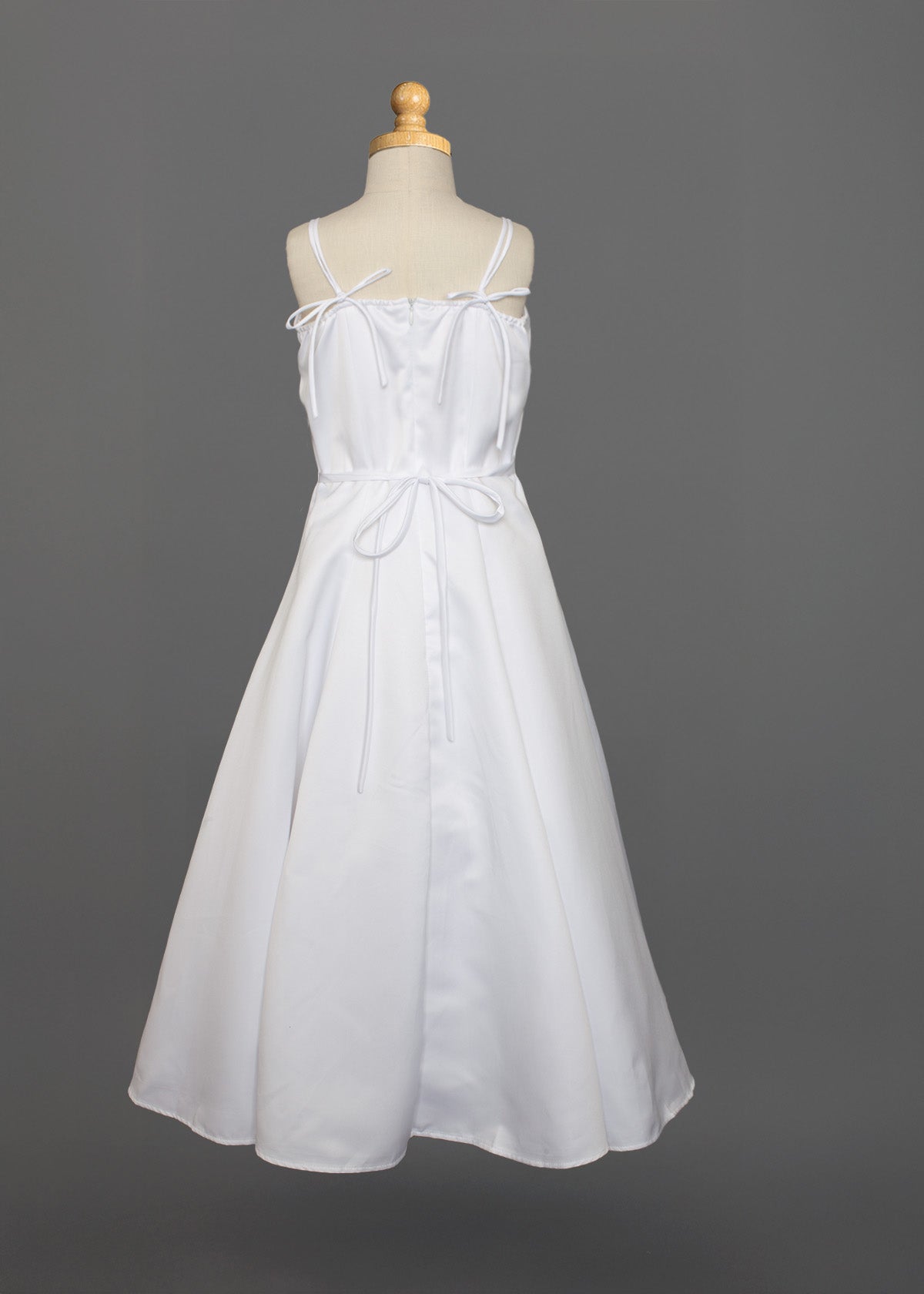 The Mabel and Honey Satin A-Line Communion Dress features hand-beaded embellishments, a sleeveless white design with thin straps, two bows at the chest and waist, displayed ankle-length on a dress form against a gray background.