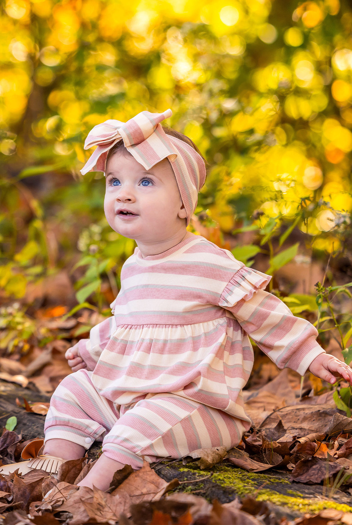 A baby wearing the Mabel and Honey Candy Stripe Romper with ruffled shoulders and a matching headband sits among fallen leaves, surrounded by yellow and green foliage, looking up and smiling.