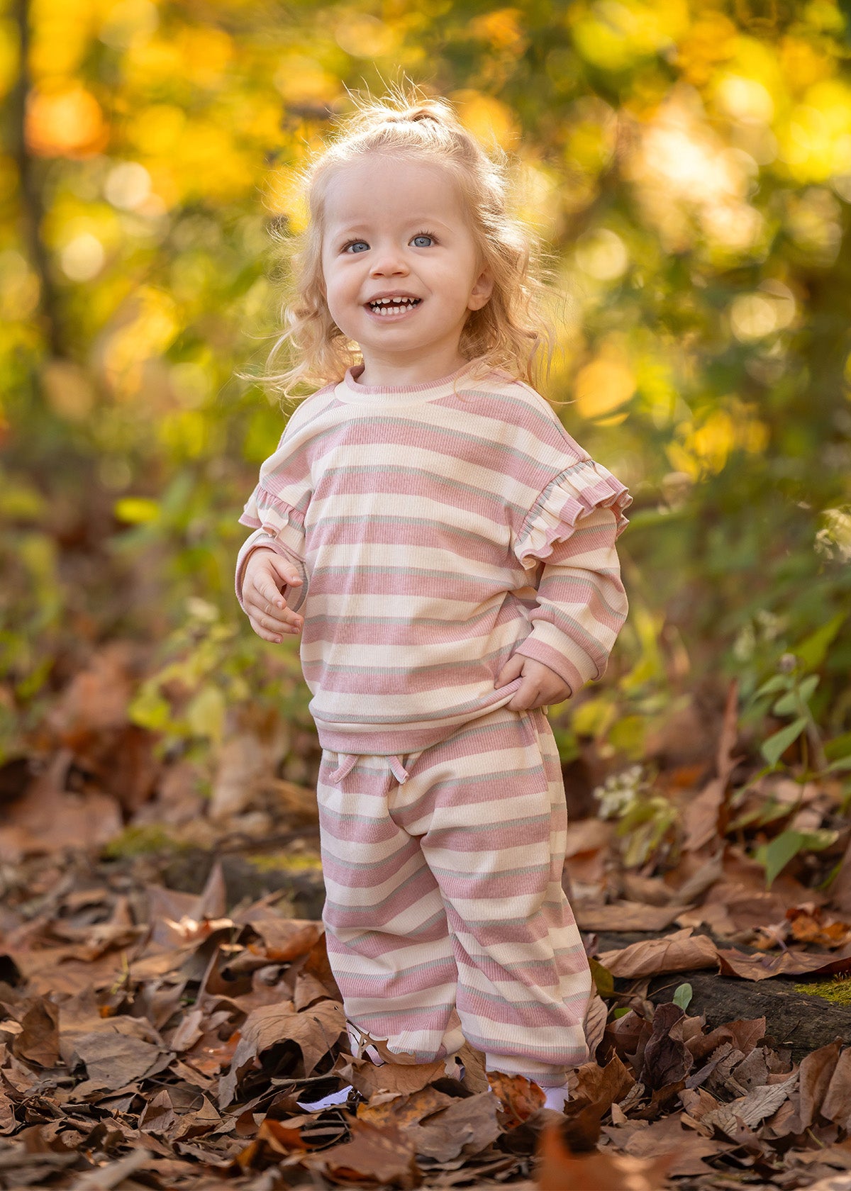 A smiling toddler with curly blonde hair stands among autumn leaves, wearing the cozy Mabel and Honey Candy Stripe Two Piece Set with ruffled sleeves that adds playful charm to her outdoor style.