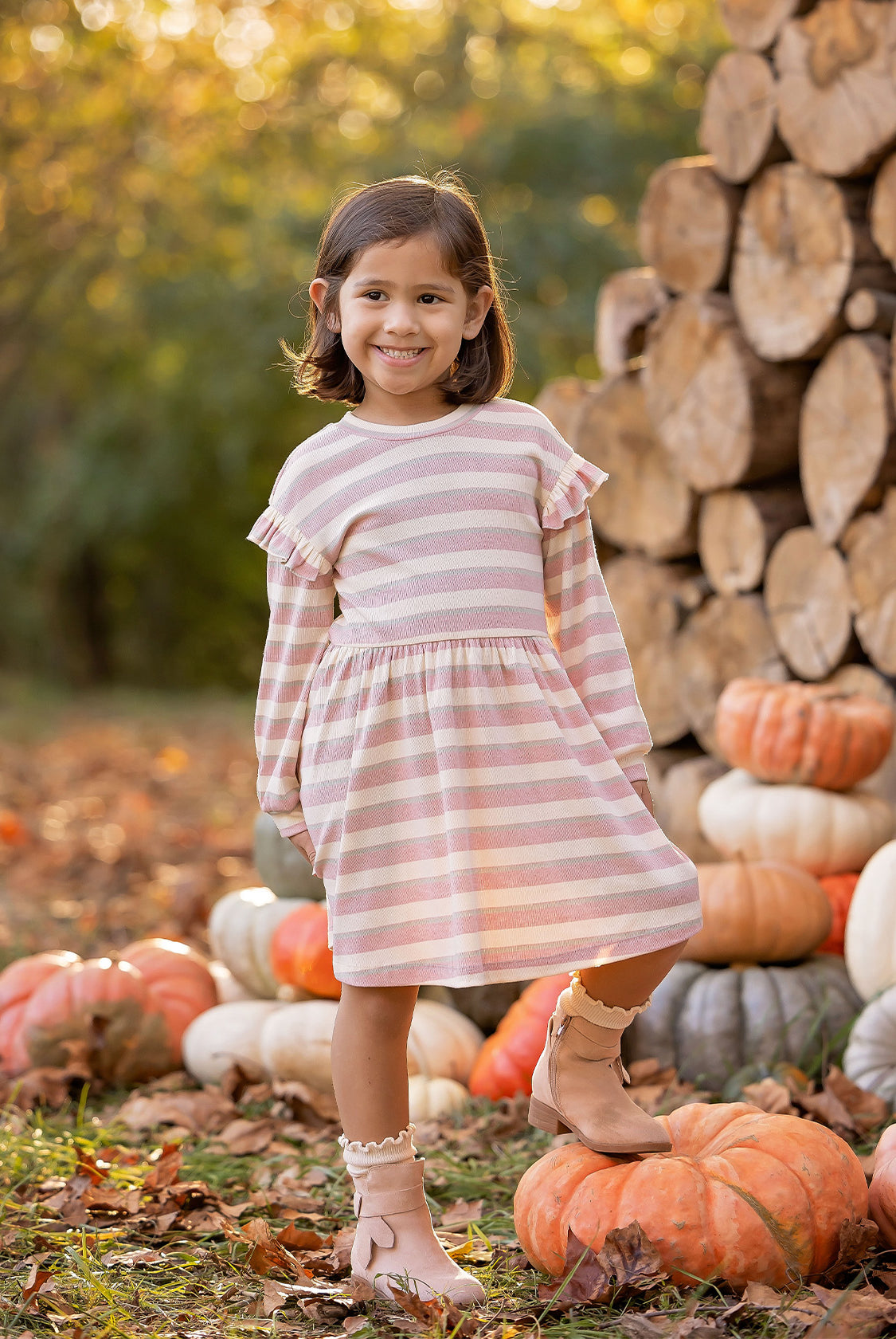 A young girl beams outdoors in the Mabel and Honey Candy Stripe Dress, surrounded by stacked logs and pumpkins with autumn trees behind her—perfectly capturing the essence of fall.