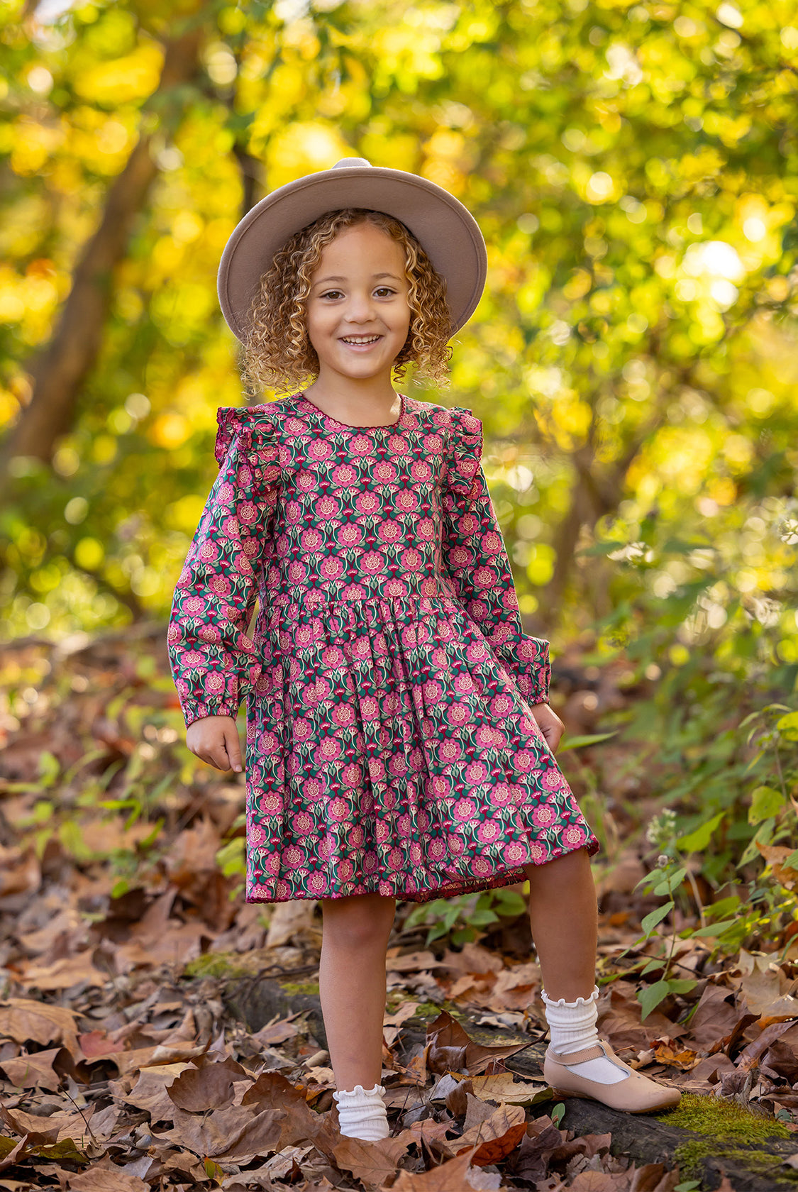 A young girl with curly hair smiles outdoors on fallen leaves, wearing the Mabel and Honey Perfect in Pink Dress with ruffle details, a wide-brim hat, white socks, and tan shoes. Sunlight filters through the trees behind her.