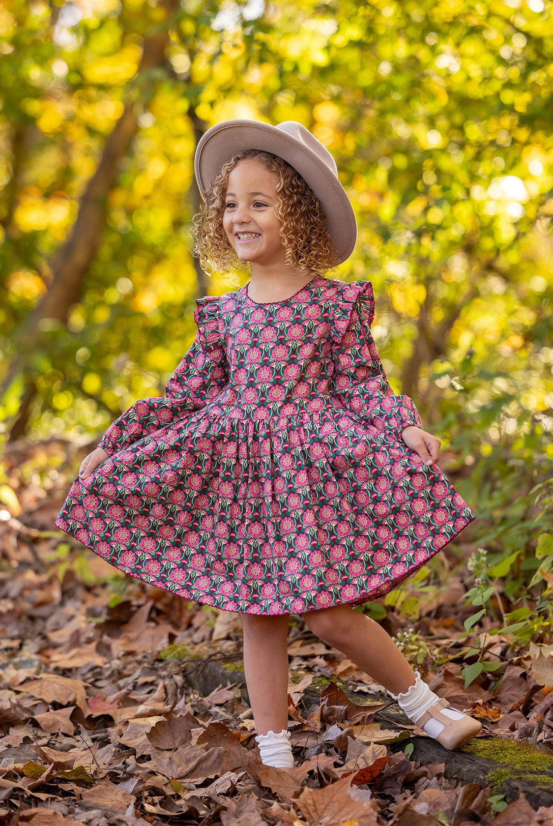 A young child smiles outdoors, wearing the Mabel and Honey Perfect in Pink Dress with ruffle details, white socks, brown shoes, and a light hat, standing among fallen leaves in a sunlit woodland setting.