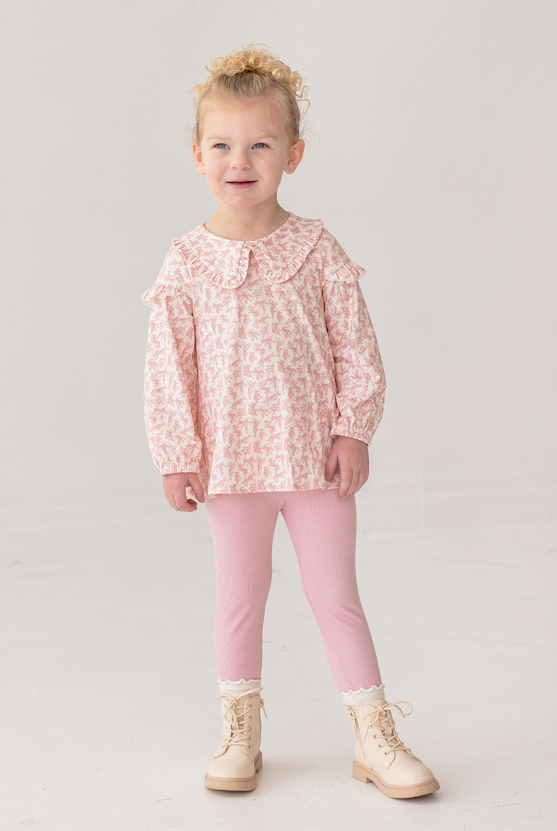 A young child smiles in the Mabel and Honey Primrose Two Piece Set—featuring a ruffled, long-sleeved floral top and pink leggings—paired with cream lace-up boots against a plain light background.