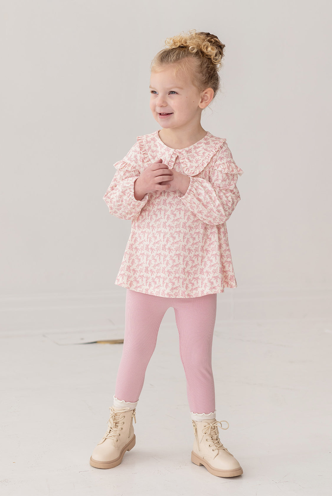 A young girl with curly blonde hair in a bun smiles while wearing the Mabel and Honey Primrose Two Piece Set—featuring a floral top and matching pink leggings—an adorable baby girl outfit, set against a plain white background.