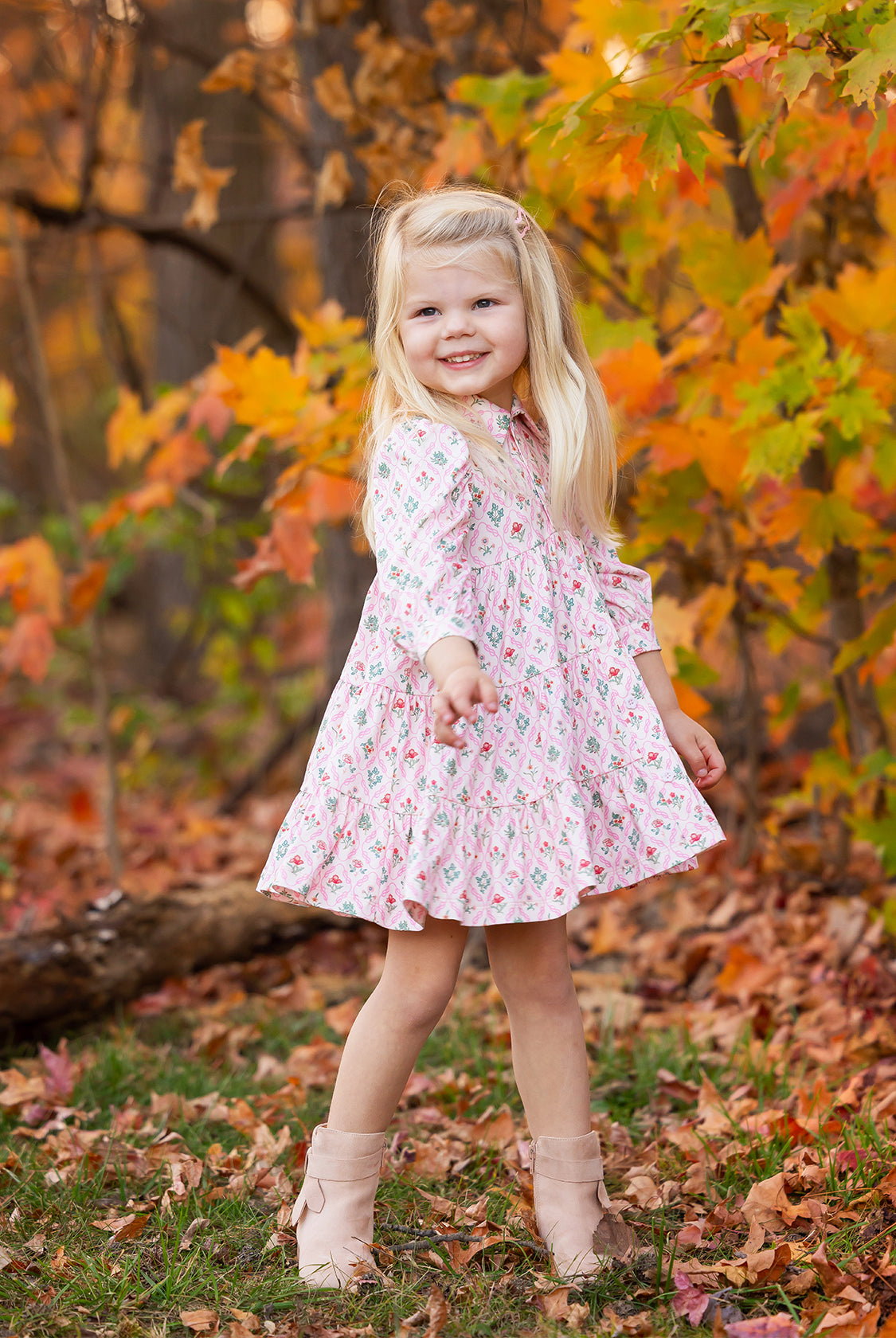 A young girl with long blonde hair smiles outdoors among autumn leaves, wearing the Dixie Pink Floral Dress by Mabel and Honey with a tiered silhouette and light boots.