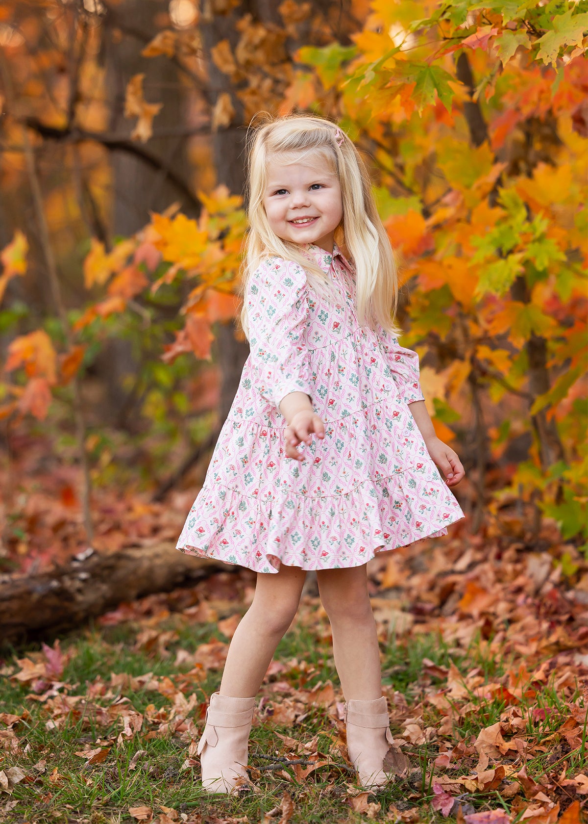 A young girl with long blonde hair smiles outdoors among autumn leaves, wearing the Dixie Pink Floral Dress by Mabel and Honey with a tiered silhouette and light boots.