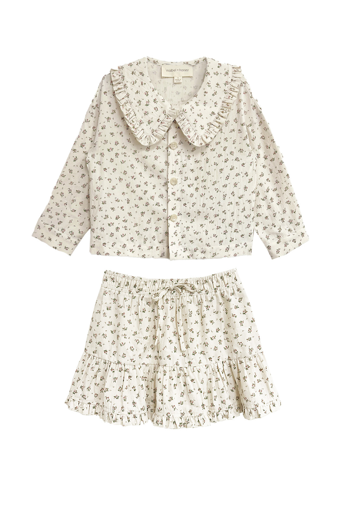 The Juliette Two Piece Set by Mabel and Honey is a charming girls’ outfit with a ruffled-collar button-up top and drawstring skirt, both in lightweight fabric with a delicate floral print.