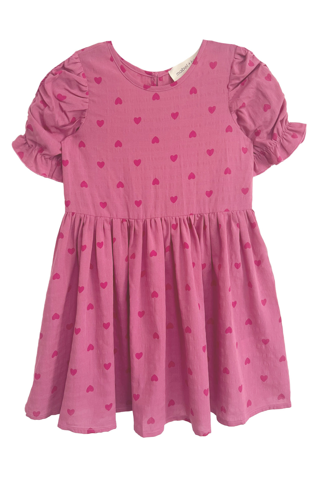 The Mabel and Honey Perfect in Pink Hearts Short Sleeve Dress is a girls’ pink dress with puffed ruffled sleeves, a flared skirt, and a sweet pattern of small, darker pink hearts.